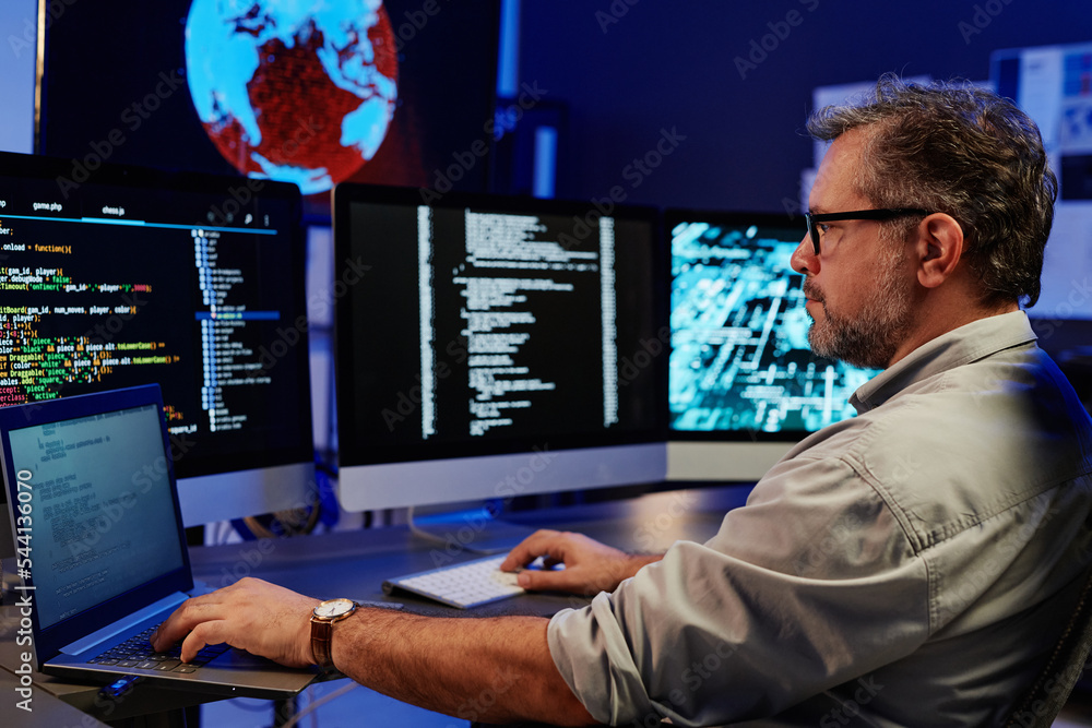 Poster Side view of serious server engineer using laptop and computers while sitting in front of monitors by workplace and decoding data