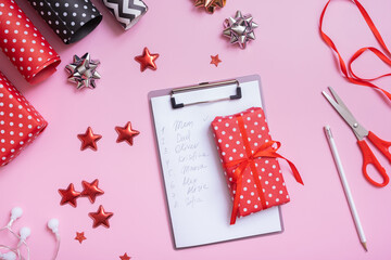 List of names and Christmas gift with wrapping materials flat lay on pink background. Top view of...