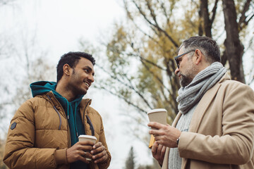 Friends, a senior and a young man walking and talking and drinking coffee together in the autumn park..