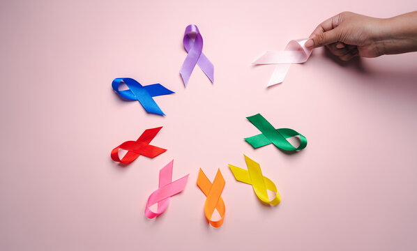 Hand holding pink ribbon which is among colorful ribbons on pink background, cancer awareness, World cancer day, National cancer survivor day, world autism, supported living and illness