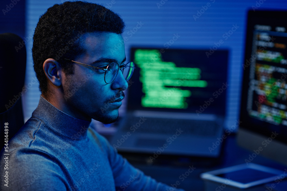 Poster Young serious IT support engineer in eyeglasses looking at computer screen while sitting in front of monitors and decoding data