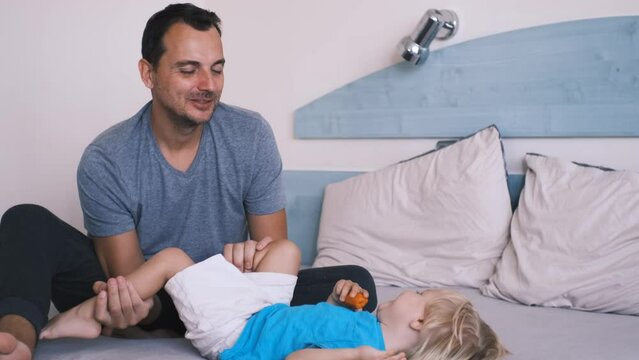 father plays with his little son on the bed, kisses his leg and they laugh