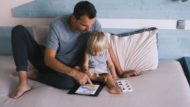 A small caucasian boy with his father on the bed draw with colored pencils.