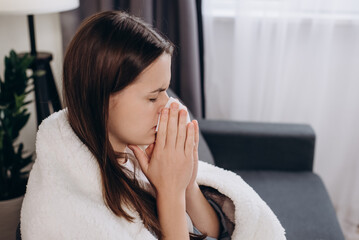 Close up of sick young woman sit on couch covered in warm plaid, suffering from flu, treatment at home. Unhealthy brunette female blowing nose, coughing or sneezing in tissue. Cold and fever concept