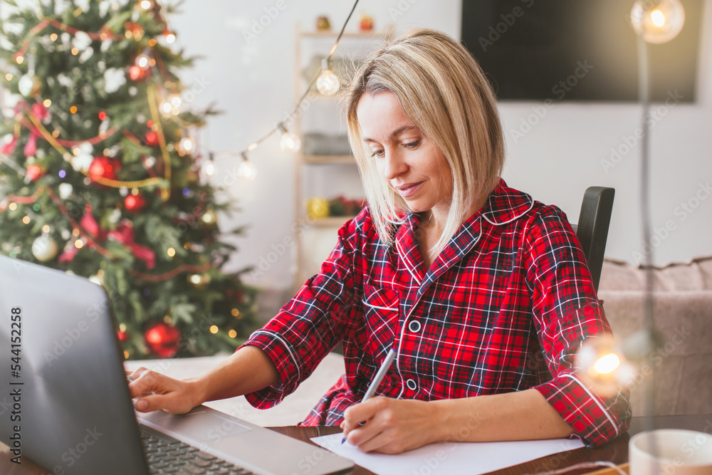 Wall mural Middle aged woman works at home remotely with laptop on the background of Christmas tree