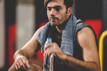 Handsome young Asian man sitting on modern gym and club floor with a towel on neck drinking water and taking rest and break after a heavy routine of workout for a fit and healthy lifestyle
