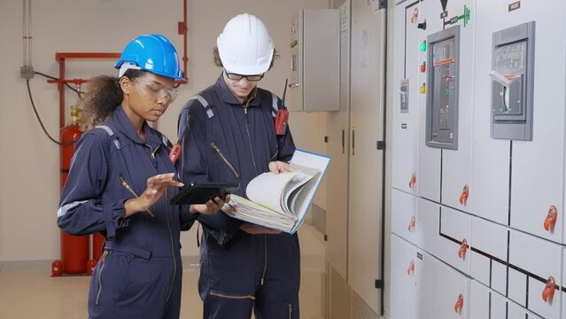 Electrical young woman and man engineer examining maintenance cabinet system electric and looking manual book in the control room at factory, technician or electrician inspection power distribution.