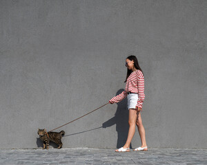 A young woman walks with a gray tabby cat on a leash against a gray wall. 