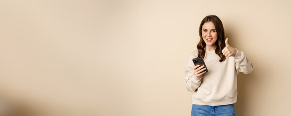 Young woman smiling, showing thumbs up while using mobile phone, smartphone app, standing over...