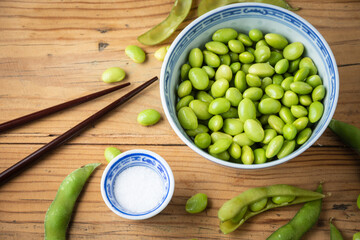 Close-up of edamame grains in blue Chinese bowl, bowl with salt on wooden table with pods, grains and chopsticks, horizontal