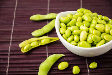 Close-up of edamame beans in white bowl on purple oriental mat with pods and beans, horizontal