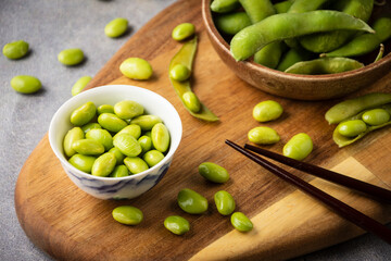 Top view of edamame beans in white bowl and pods in wooden bowl on table with pods, beans and chopsticks, horizontal