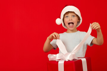 Boy opens his mouth pulling up the white ribbons of large bow on red holiday box. Boy in funny red Santa Claus cap unpacks gift box