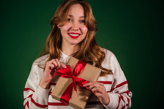 Smiling young woman with white teeth holding new year gift isolated on green background. Girl opening christmas present looking at camera
