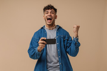 Young handsome stylish enthusiastic man holding phone doing winner gesture