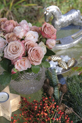 A bouquet of Cappuccino roses in a vase on the table with natural materials for Christmas decor: branches or holly holly, rose fruits of Multiflora, larch cones, wooden rocking chair.