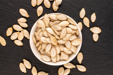 Unpeeled raw pumpkin seeds in white ceramic plate on slate stone, macro, top view.