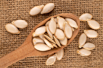 Unpeeled raw pumpkin seeds with wooden spoon on jute cloth, macro, top view.