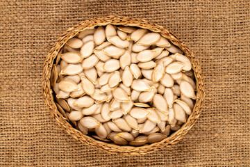 Unpeeled raw pumpkin seeds in straw plate on jute cloth, macro, top view.