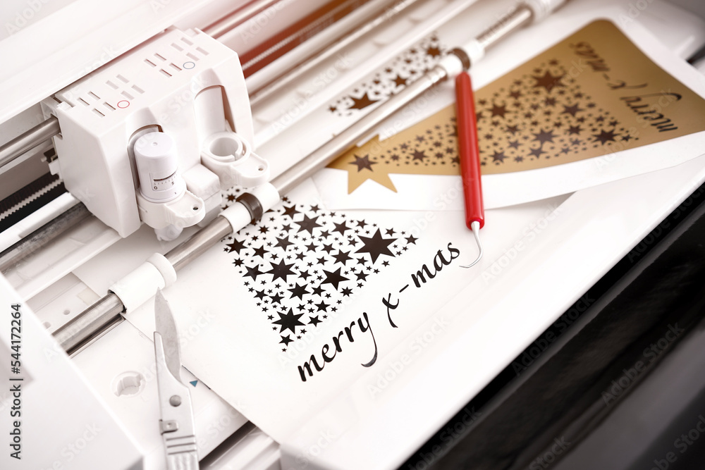 Fototapete closeup view on machine head of plotting machine that cuts christmas sticker with lettering from black adhesive vinyl. DIY concept with tools and additional sticker. warm light mood. selective focus