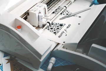 plotting machine cuts christmas sticker with stars and lettering from black vinyl. work scene with tools and additional stickers. cold misty light mood. selective focus