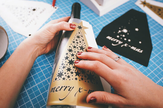 female hands with painted nails put a golden christmas ornament sticker on a wine bottle. weeding tool, scalpel and plotter blurred out in background. holiday gift and advertisement concept