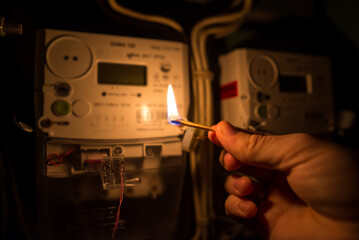 A man's hand in complete darkness holds a burning match to read the home electricity meter.
