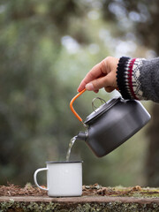 pouring hot water from metal teapot into a enamel vintage cup at wooden table outdoors in autumn-winter forest