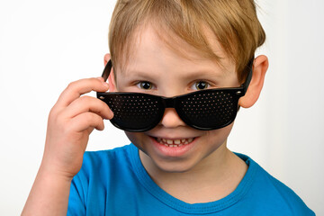 A little boy in black medical glasses for vision correction on a white background. He holds his glasses with his hand and smiles
