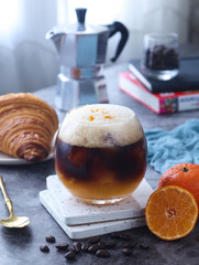 A clear glass of iced-orange americano coffee topped with zest of orange. Croissant and moka pot in...