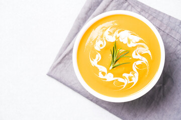 Thick butternut squash bisque in white bowl garnished with heavy cream and rosemary over white table. Bowl of pumpkin cream soup. View overhead. Minimal style. Selective focus