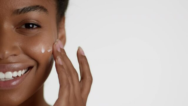 Premium facial cosmetics. Half face portrait of young happy african american woman rubbing cream on cheek, empty space