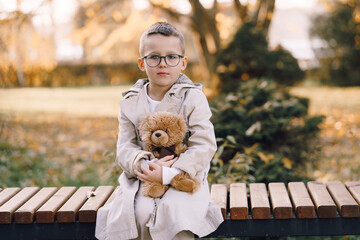 Little boy wearing a beige raincoat holds a teddy bear in his hands. Little kid are playing in park. Warm autumn.