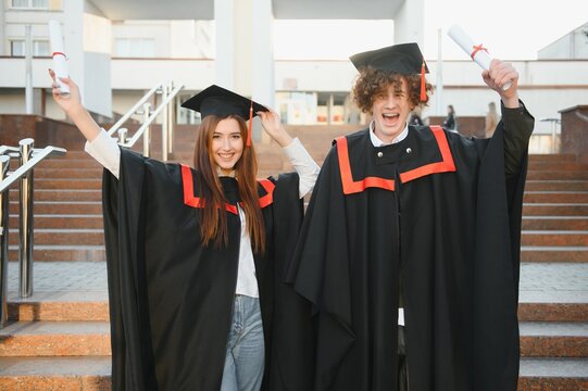 Portrait of happy graduates. Two friends in graduation caps and gowns standing outside university building with other students in background, holding diploma scrolls, and smiling