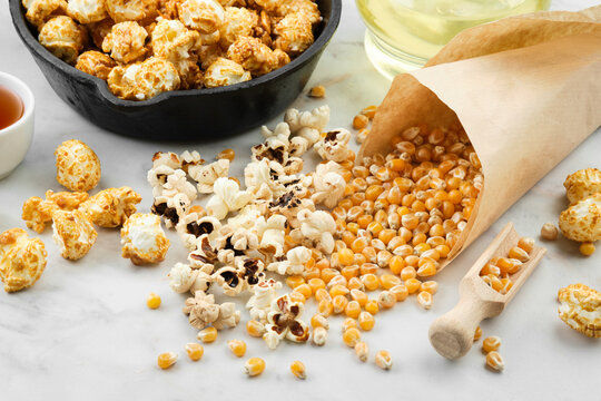 Prepared popcorn in a frying pan, dried corn grains, caramel sauce and oil bottle on kitchen table.