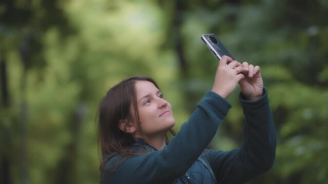 A young woman taking pictures of the trees with a phone, blurred green natural background. Slow motion. 