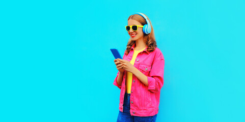 Portrait of stylish modern happy smiling young woman in headphones listening to music with smartphone wearing pink jacket on vivid blue background