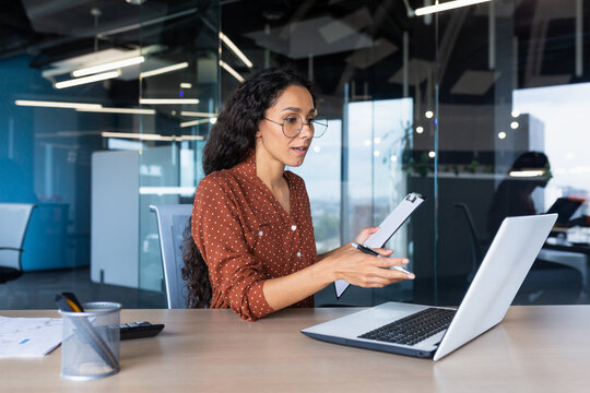 Portrait successful businesswoman, Latin American woman shows a report of financial documents online interlocutors in the laptop screen, an employee inside an office building online video conference. - Powered by Adobe