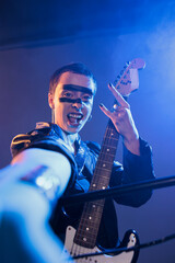 Obraz premium Crazy performer playing guitar in studio with smoke, showing rock sign with fingers and acting stylish. Cool ecstatic rocker creating live performance with heavy metal and punk music.