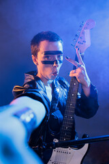Obraz premium Female artist showing peace sign on camera, singing rock music and heavy metal alternative audio. Playing guitar and doing live performance in studio, doing victory symbol with instrument.