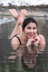 Plus size model in bathing suit lying down in geothermal mineral water in outdoor pool at balneotherapy health spa, hot springs resort having balneological properties. Woman looking at camera, smiling - obrazy, fototapety, plakaty