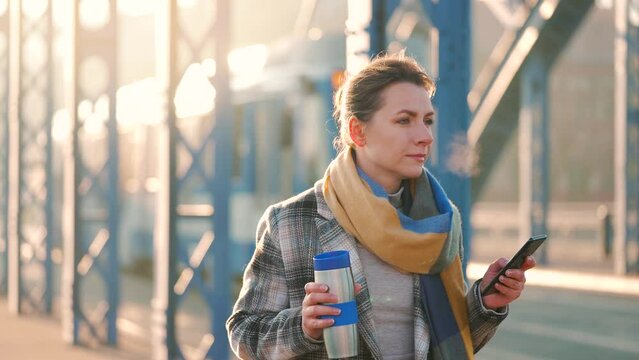 Woman walking in a frosty sunny morning, drinking coffee and using smartphone