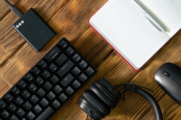 Black computer keyboard, external ssd, headphones, notepad with pen and computer mouse on a table made of wooden pine boards. Eco office. Periphery for gadgets and computer.