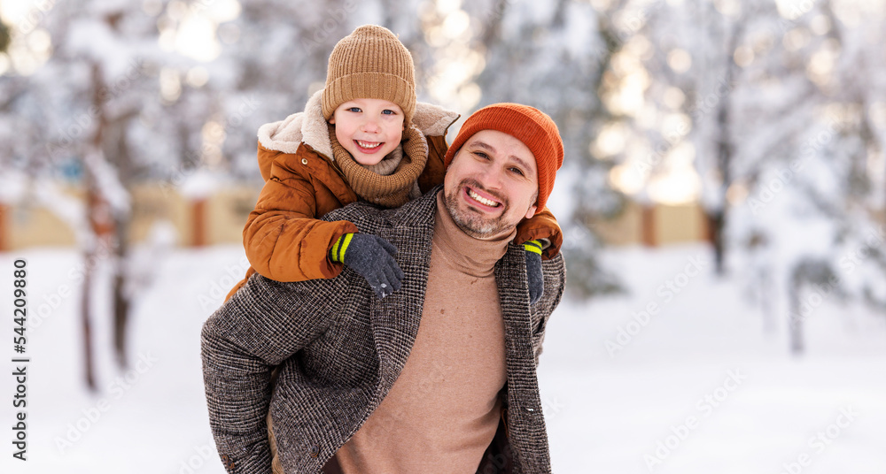 Poster dad giving piggyback ride to happy little son while walking outdoors in snowy winter park