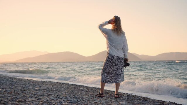 Professional photographer blonde young woman near sea on shore. sunset, unrecognizable girl photographs nature and ocean waves at dawn. traveler captures moments of nature while traveling.