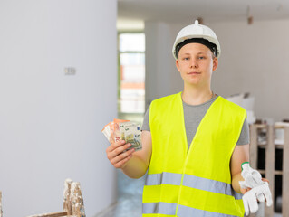 Satisfied young man in vest and helmet holding bunch of cash he obtained for first part-time job in building site. Boy got his first wage.