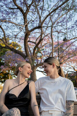 Fototapeta premium lesbian couple sitting on park bench under beautiful purple tree laughing at eachother 