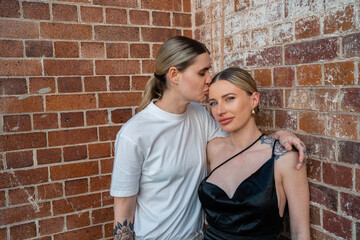 cool lesbian couple kiss on forehead in front of brick wall 