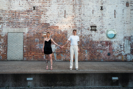 lesbian couple holding hands in front of brick wall looking at camera