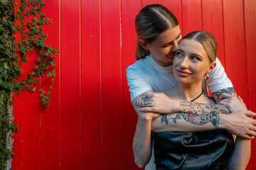 lesbian couple hugging from behind in front of red wall 